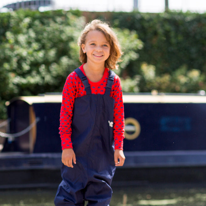 A young girl smiles wearing navy Originals Waterproof Dungarees, standing outdoors near a canal barge and lush greenery.